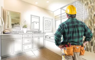 Man in yellow hard had and green plaid shirt with work belt seen from behind looking at a schematic drawing of a bathroom