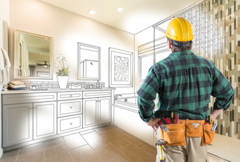 Man in yellow hard had and green plaid shirt with work belt seen from behind looking at a schematic drawing of a bathroom