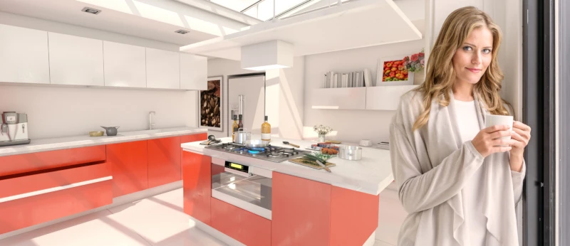 Brightly lit modern kitchen with white counters and orange cabinets. A woman stands in it with a cup of coffee.