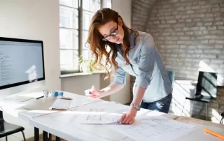 Woman with light skin and long dark hair leaning over a table looking at blueprints