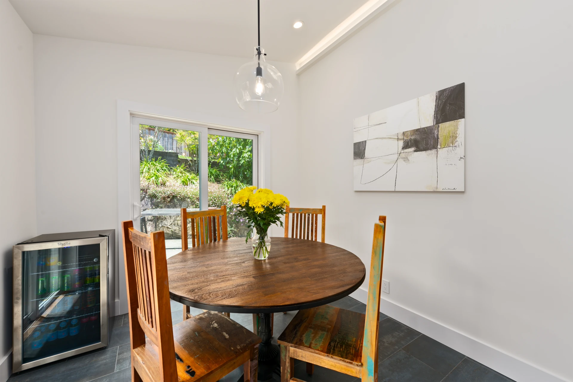 Dining area with round, natural wood table and Shaker-style chairs, modern glass pendant light, large windows and a stainless steel beverage refrigerator.