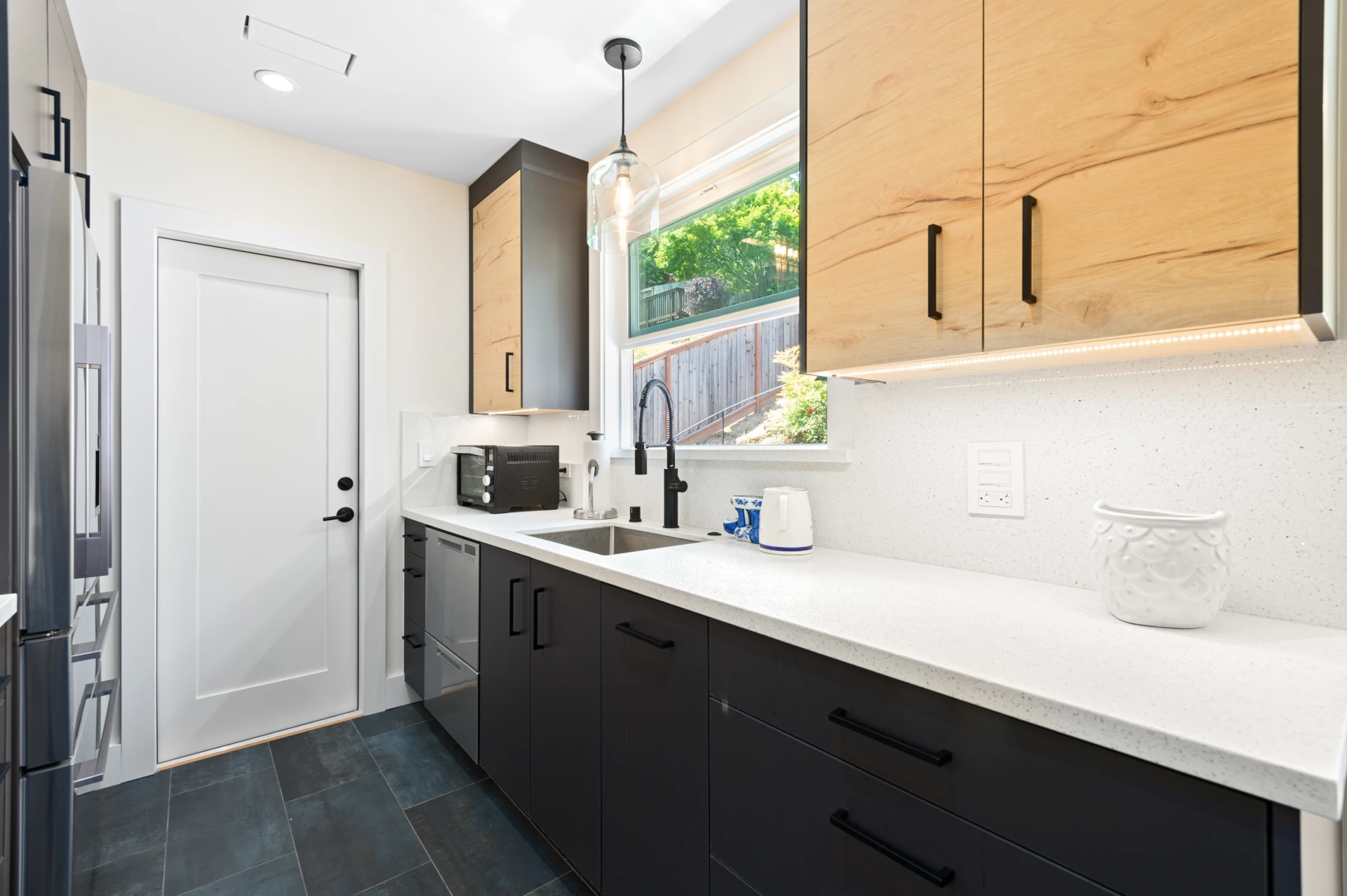 Kitchen with dark wood bottom cabinets, light wood upper cabinets, stainless-steel sink and dark commercial fixtures, stainless steel dishwasher, white quartz countertop and grey-green tile flooring.