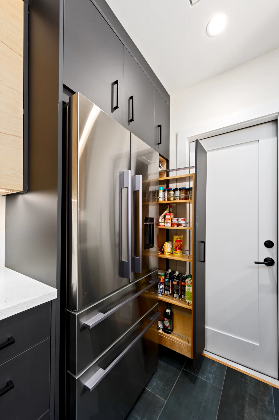 Close-up of kitchen with dark cabinets and white countertop, stainless steel French-door refrigerator an narrow pull-out spice shelf