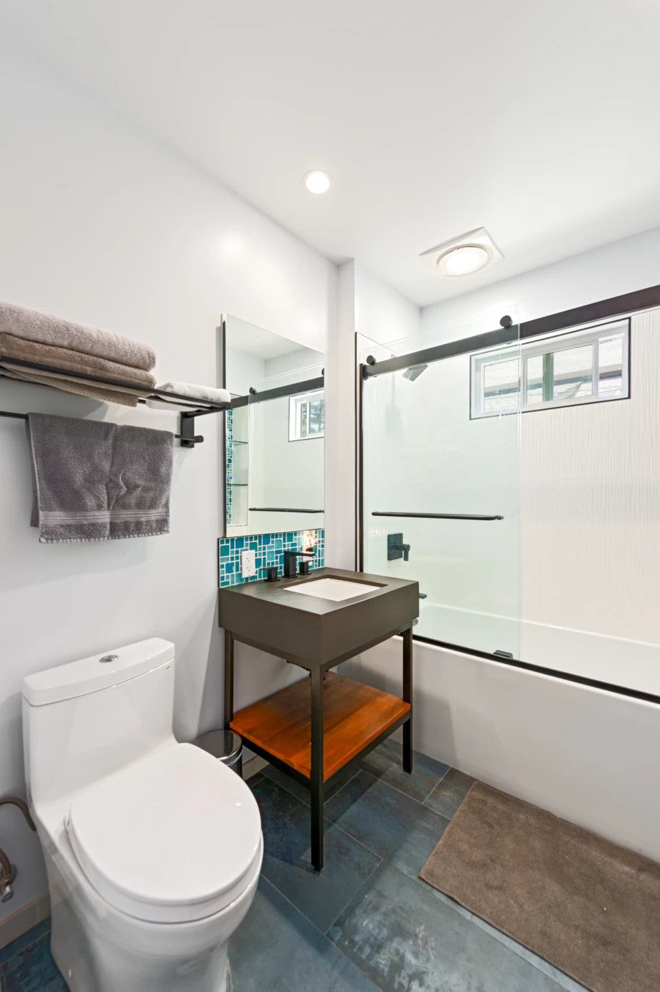 Contemporary bathroom with white tub and shower enclosure, dark wood pedastal sink with blue-green tiled backsplash, white toilet, tiled floor and brown bathmat.