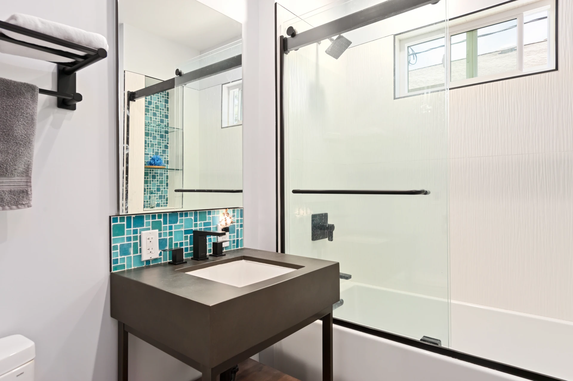 Contemporary bath with tub and shower enclosure, pedastal sink with dark wood and black modern fixtures and blue-green tile backsplash.