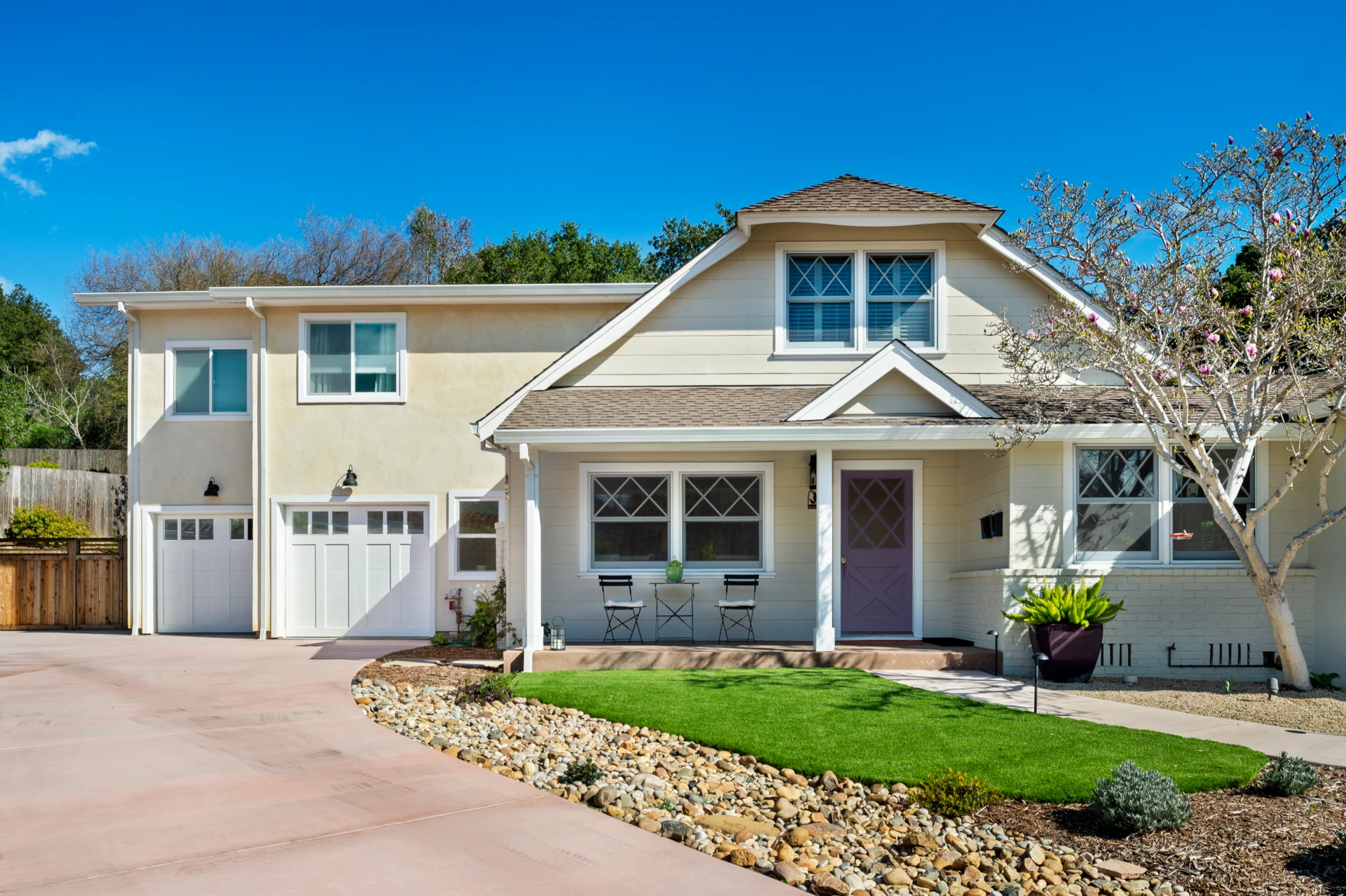 Cape-style home exterior with gabled windows and front garden with stones, green bushes, and a tree.