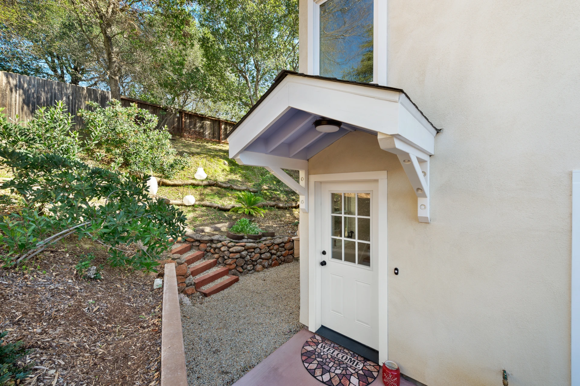 Exterior door with window topped by a gable in a beige wall with a welcome mat in front of it.