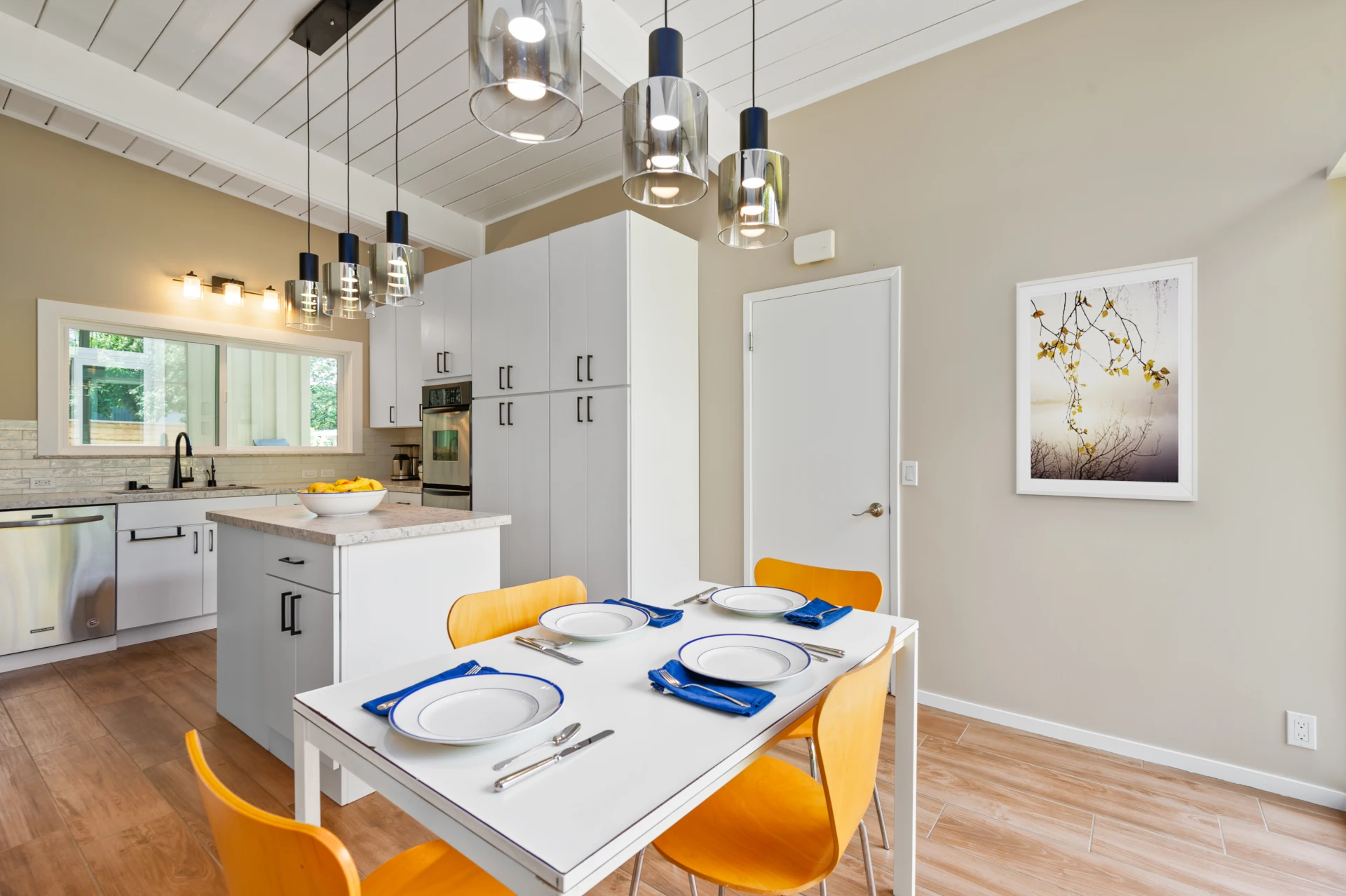 Modern kitchen and dining area with white cabinetry, beige walls, white quartz countertops, stainless-steel dishwasher and double oven, wood floors and a white rectangular table with four wood chairs.
