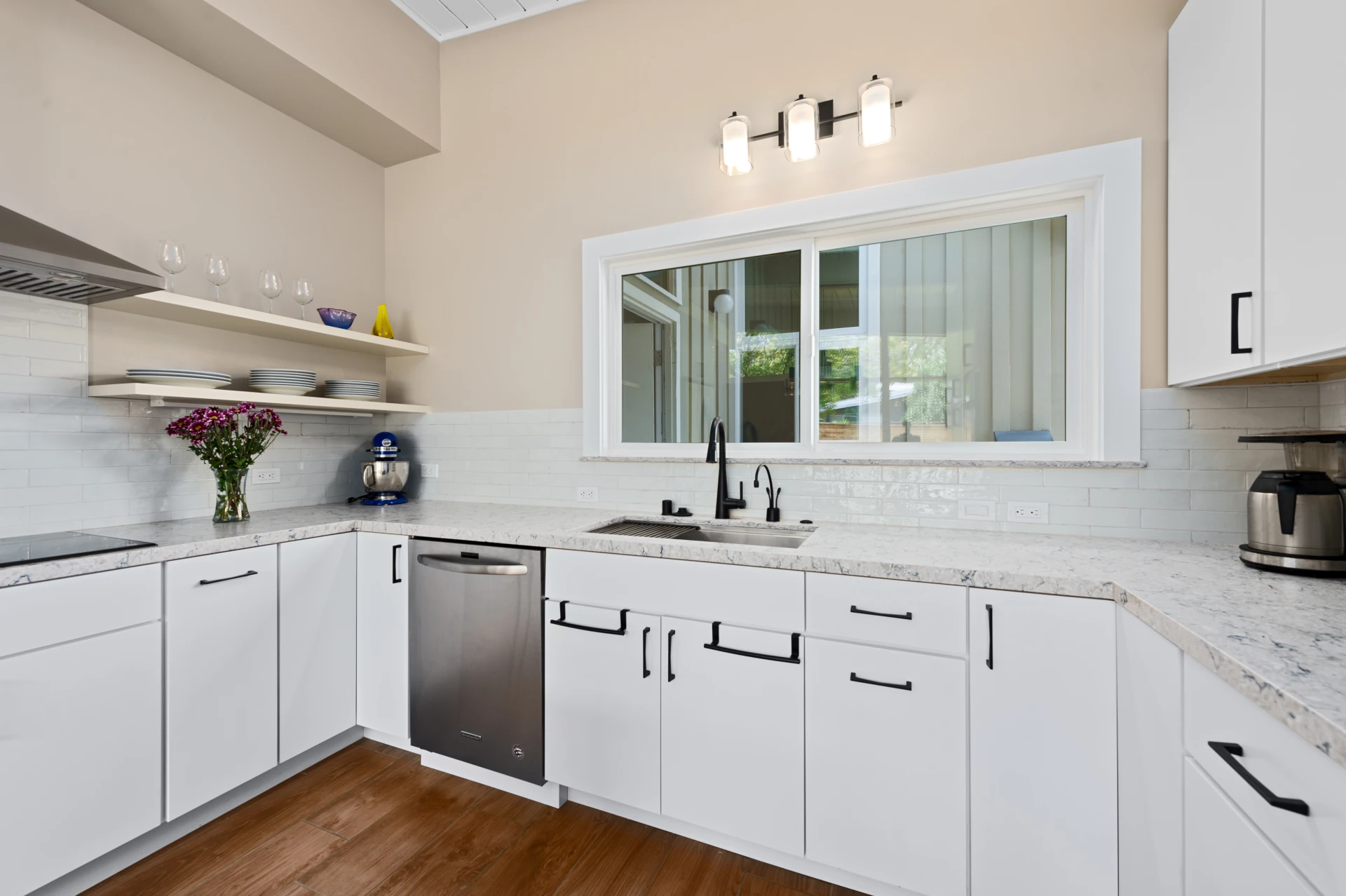 Modern kitchen with white cabinetry, beige walls, white quartz countertops, glass stovetop, stainless-steel and dishwasher and sink, with large window, and two light wood open shelves holding dishware flanking a stainless pyramid range hood. S