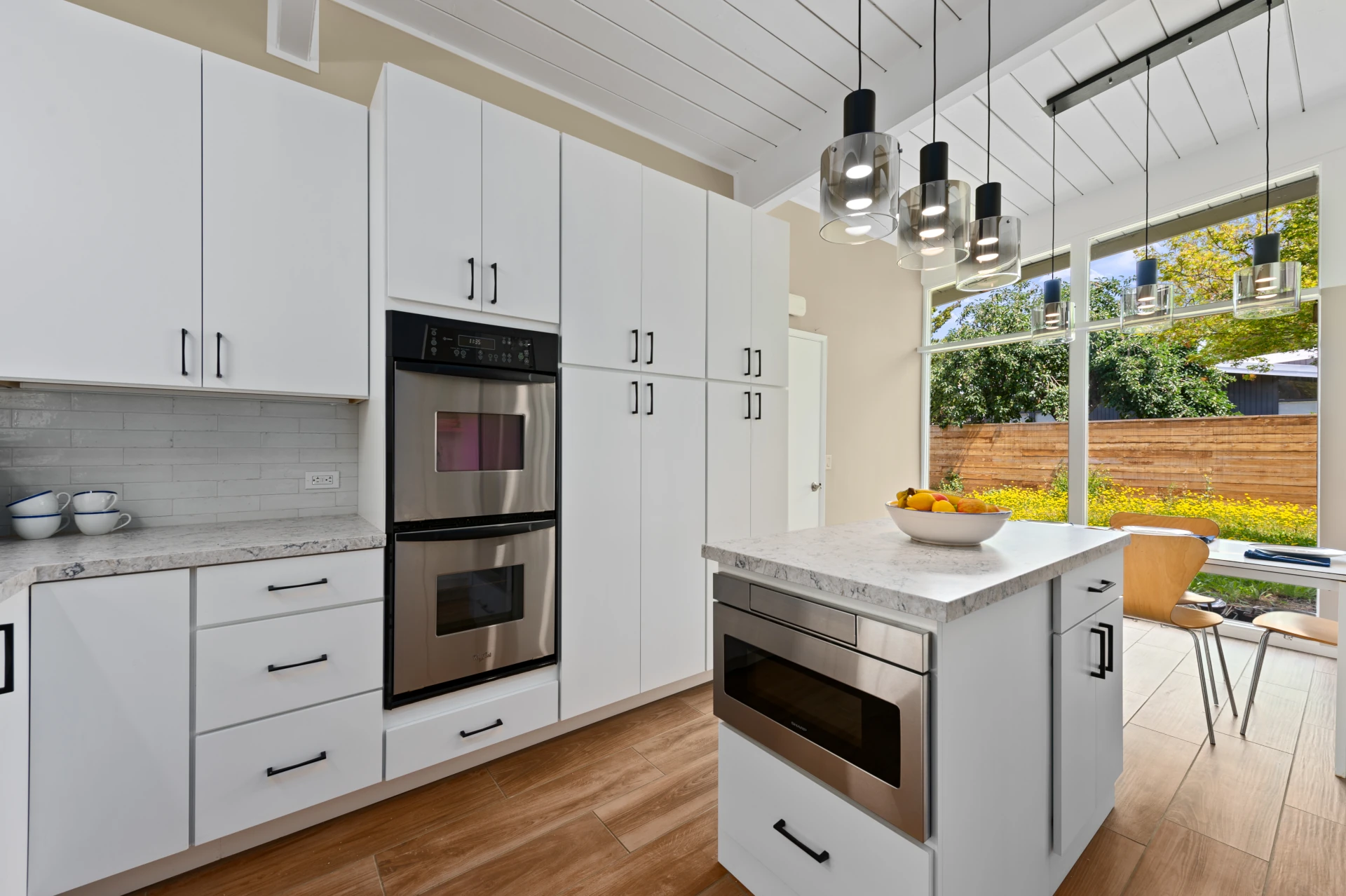 Modern kitchen and dining area with white cabinetry, beige walls, white quartz countertops, stainless-steel double oven, white island with white quartz countertop and built-in stainless microwave drawer, with glass pendant lighting and floor-to-ceiling windows opening on garden.