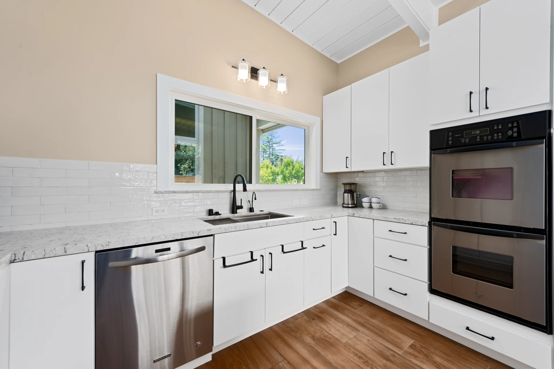 Modern kitchen and dining area with white cabinetry, beige walls, white quartz countertops, stainless-steel double oven, sink, and dishwasher, and wood floors.