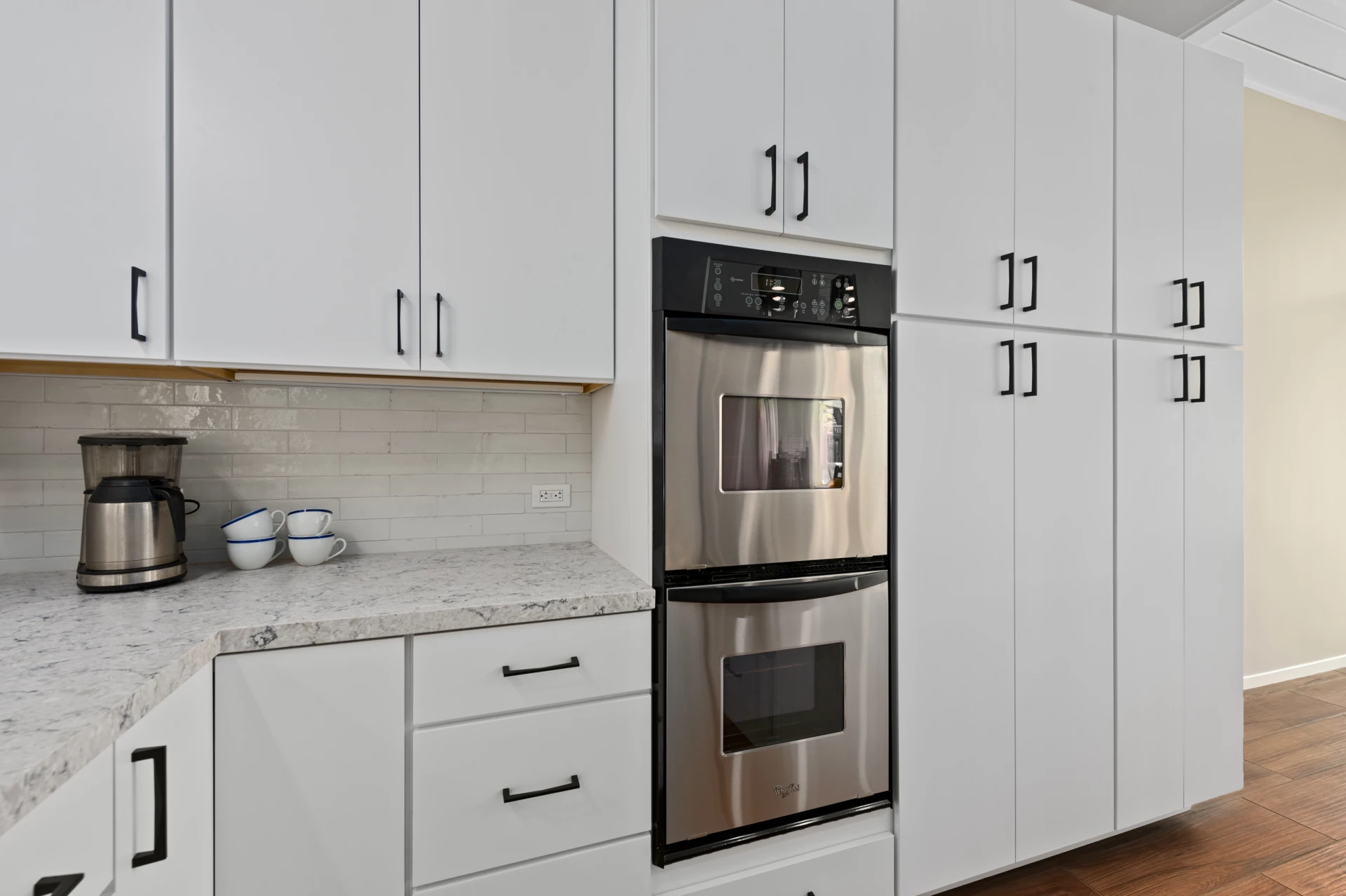 Modern kitchen and dining area with white cabinetry, beige walls, white quartz countertops, stainless-steel double oven, and wood floors.