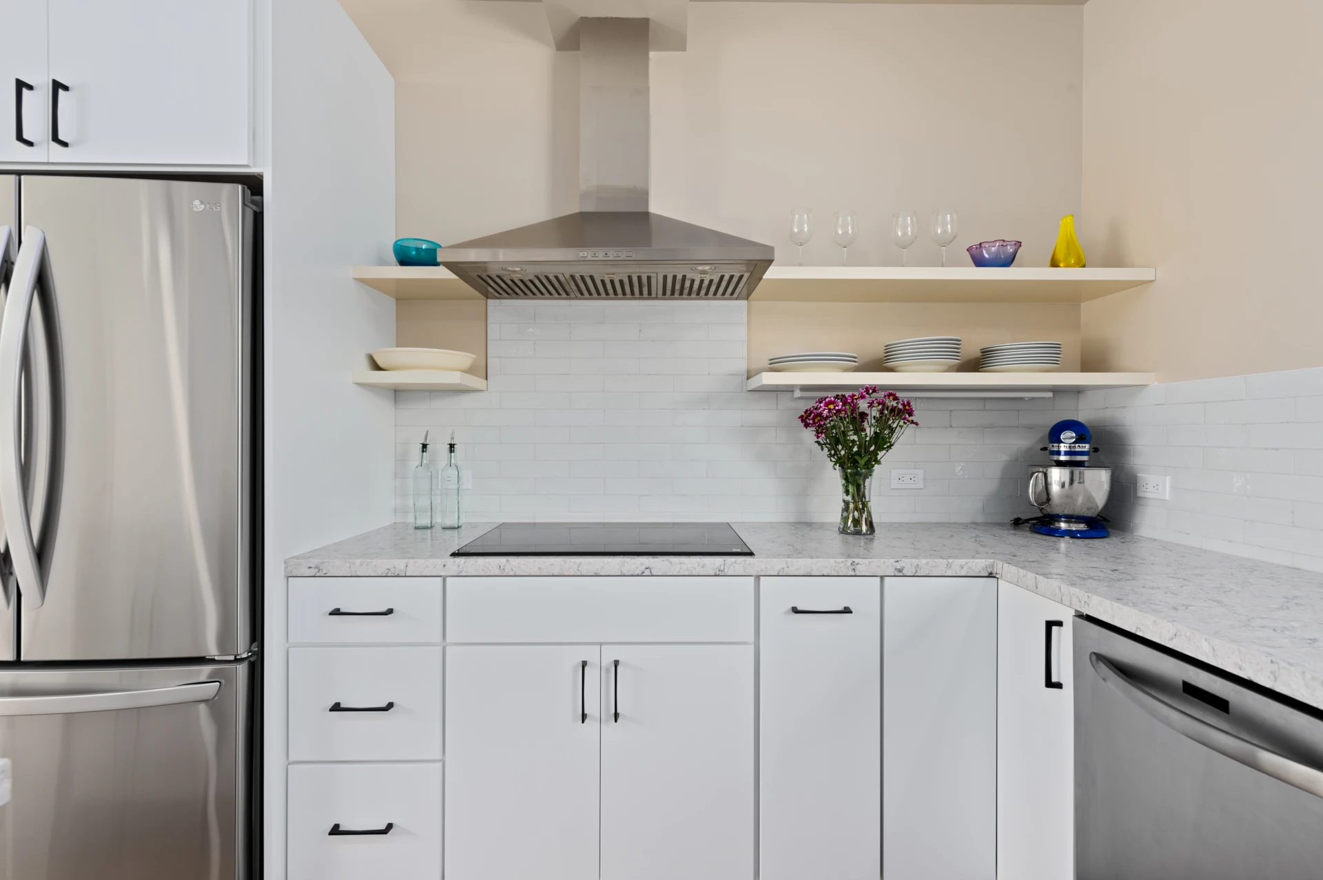 Modern kitchen with white cabinetry, beige walls, white quartz countertops, glass stovetop, stainless-steel refrigerator and dishwasher, with two light wood open shelves holding dishware flanking a stainless pyramid range hood.