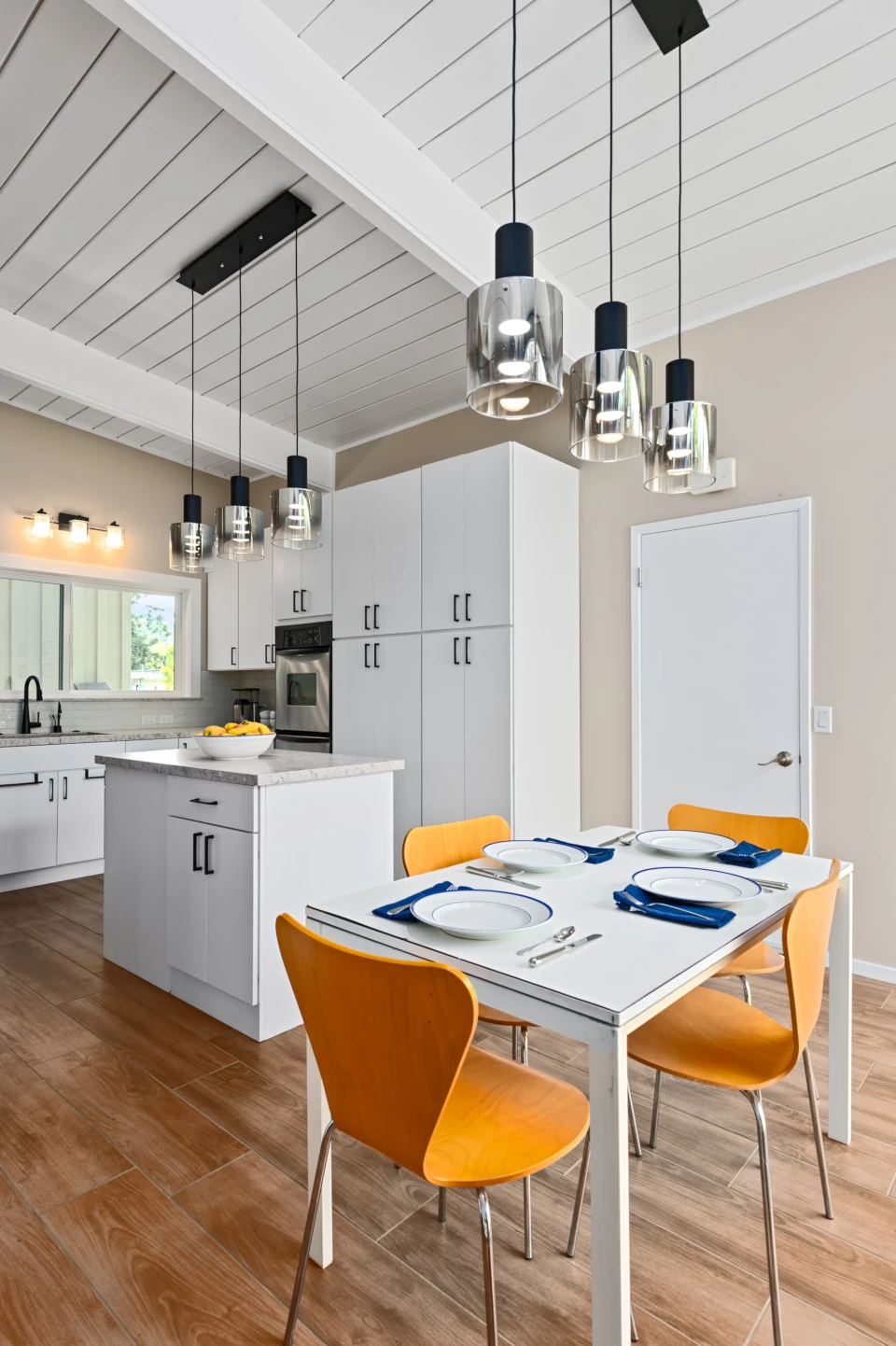 Modern kitchen and dining area with white cabinetry, beige walls, white quartz countertops, stainless-steel dishwasher and double oven, wood floors and a white rectangular table with four wood chairs. A three-pendant glass light fixture hangs above the table.