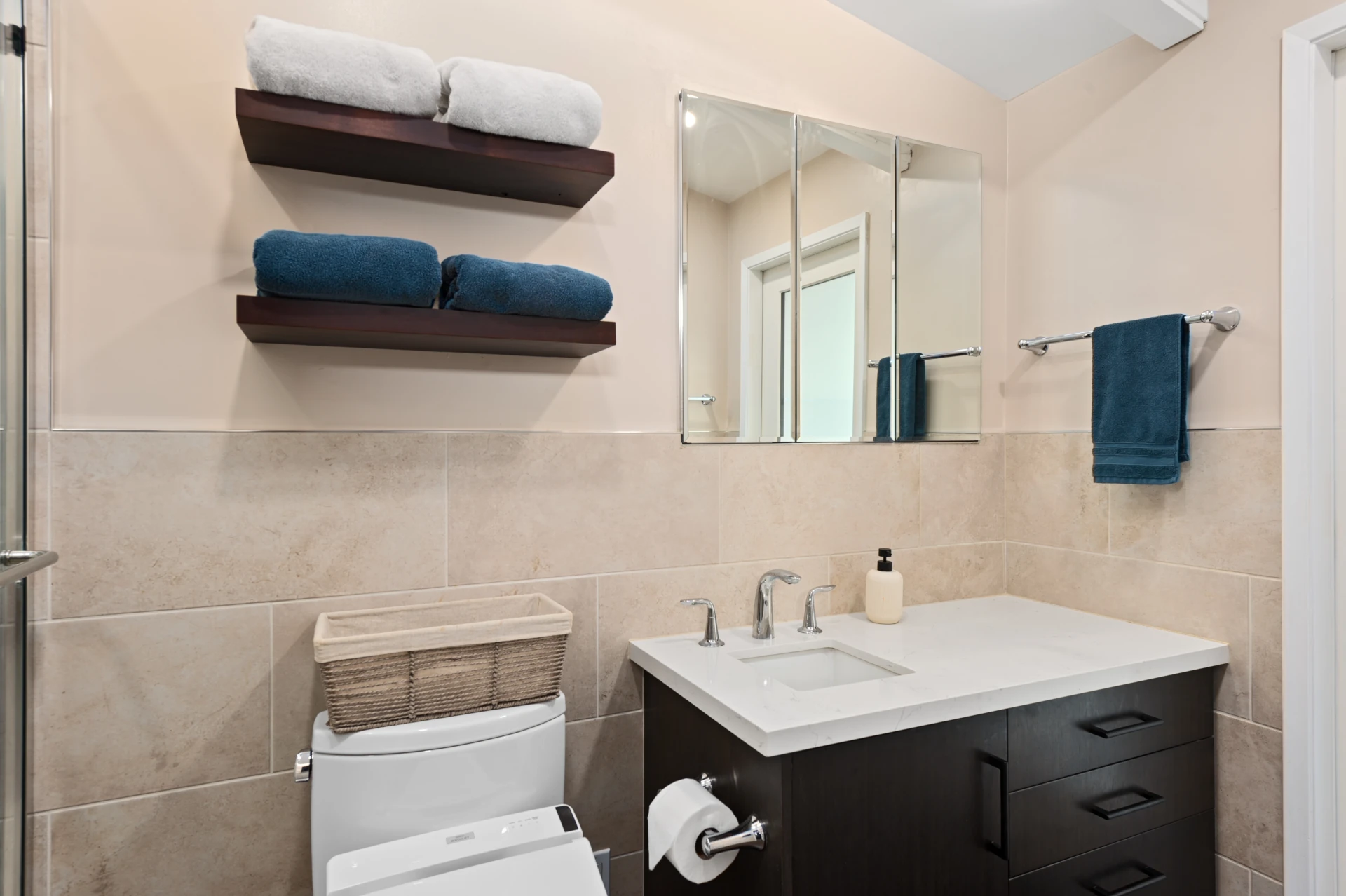 Bathroom with beige walls, a dark wood cabinet with white sink and countertop, beige ceramic tile, white toilet, and dark wood shelves with blue towels.