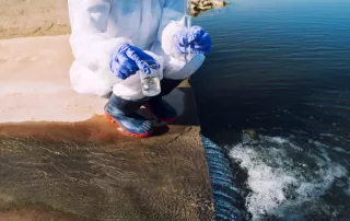 Person in white Tyvek suit kneeling beside a reservoir with a test beaker and pipette