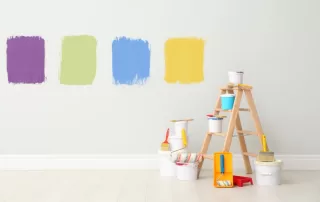 Square paint swatches of purple, green, blue, and yellow on a light-gray wall, with a small ladder and pain cans in front of it.