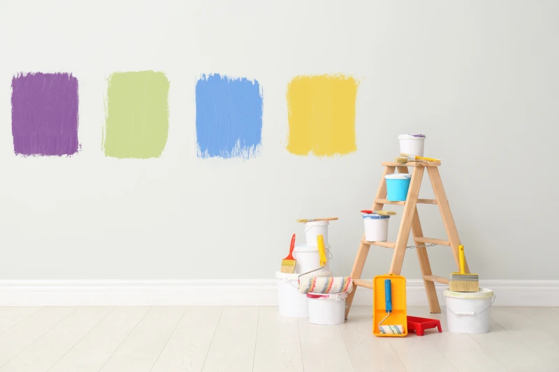 Square paint swatches of purple, green, blue, and yellow on a light-gray wall, with a small ladder and pain cans in front of it.