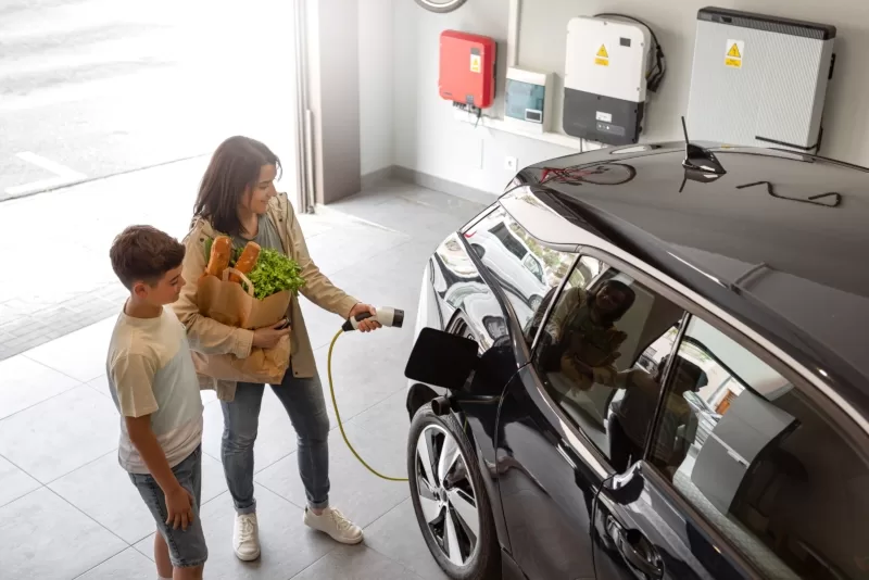 Happy son and mother plug EV charger from home charging station to electric vehicle. Eco-friendly car powered by renewable source of clean energy.